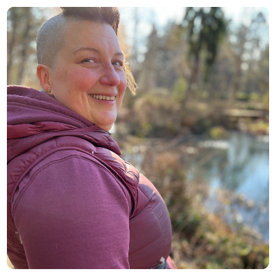 Stone is a white person with a mostly shaved head and a unicorn plume of brown hair. They are wearing a wine colored vest and hoodie, standing in profile next to a pond. 