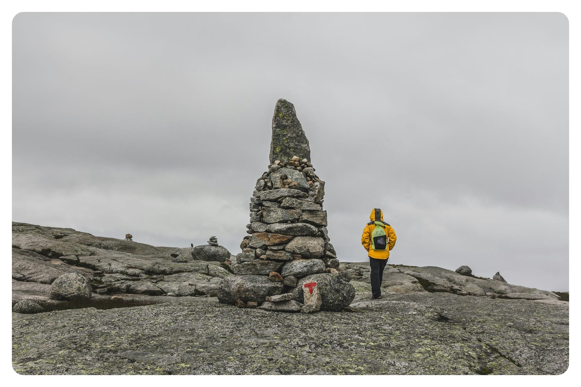 A large stone cairn marks the path across a bare, rocky landscape as a hiker in a bright yellow jacket passes by.